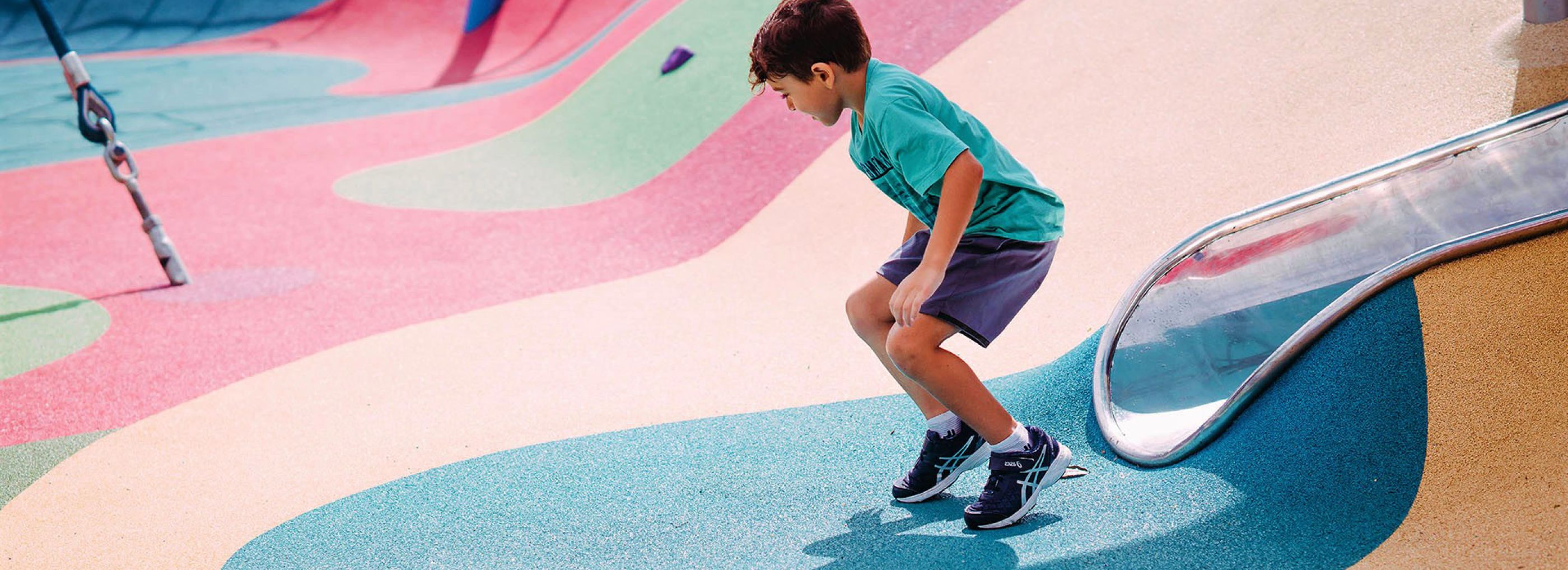 A young boy standing up after going down a slide that has been built into colourful rubber playground flooring.